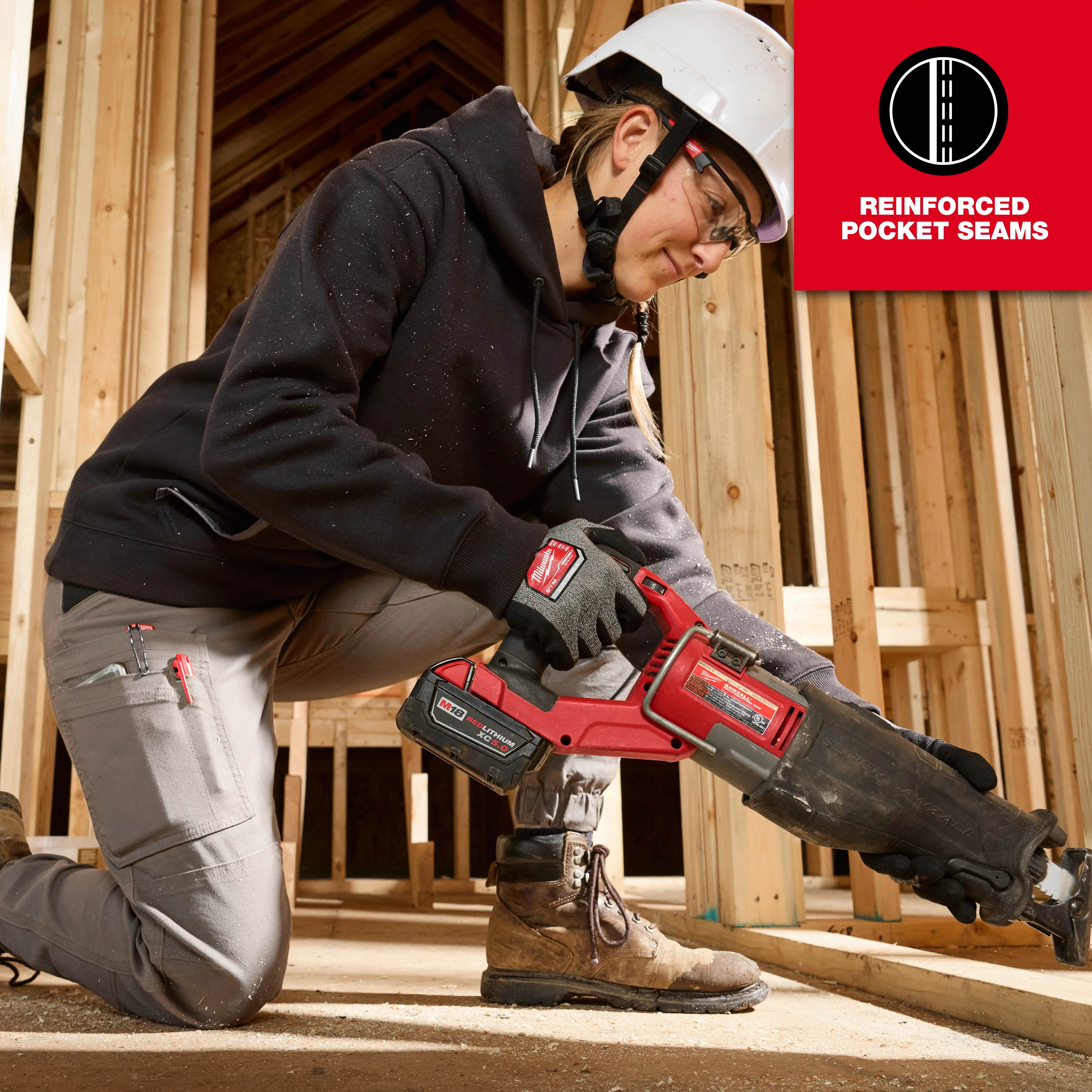 A woman wearing a black Women's FREEFLEX™ Pullover Hoodie and gray pants with reinforced pocket seams is working on a construction site, using a red power tool while kneeling. The scene is set inside a wooden framework of an unfinished building.