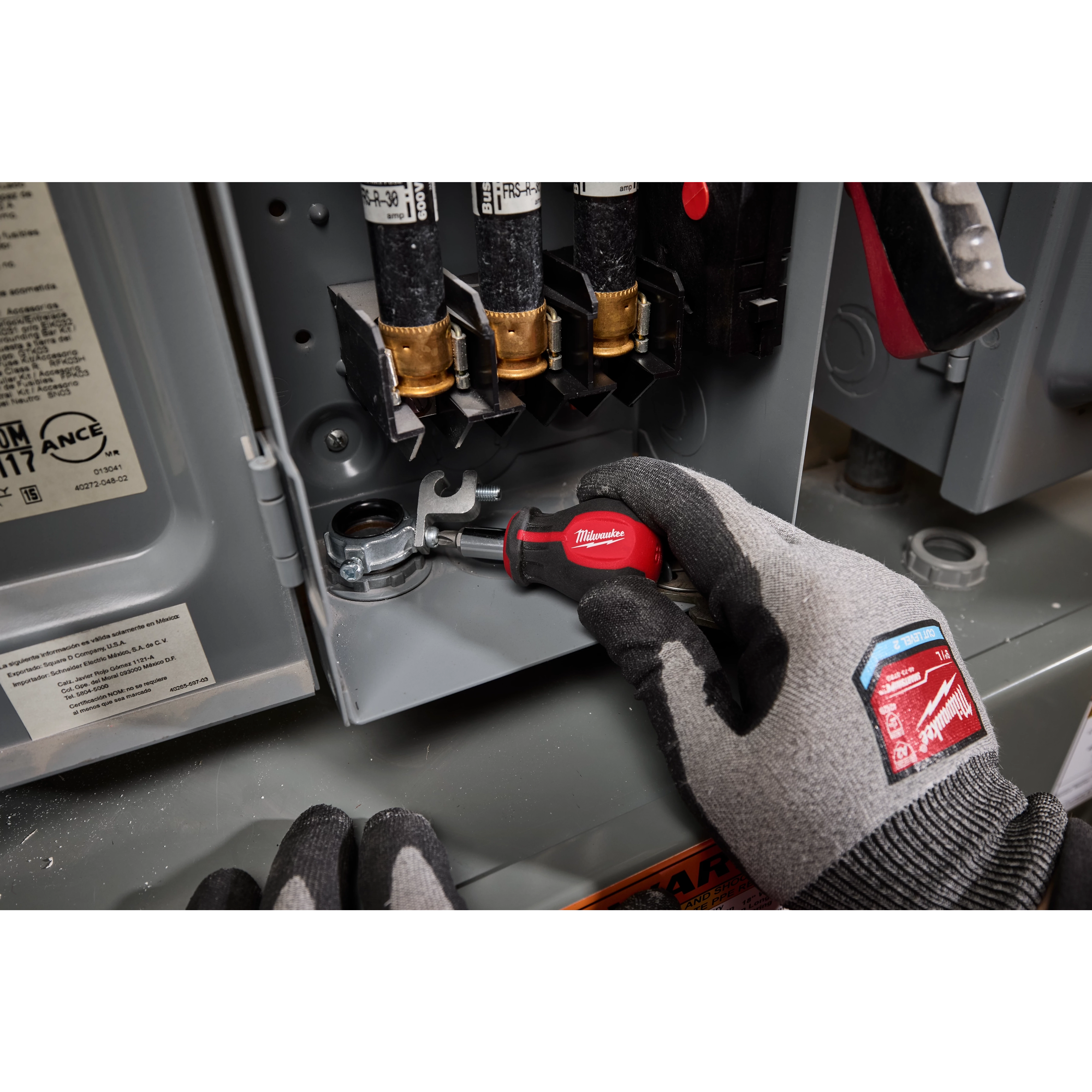 A person wearing gray gloves uses a 6-IN-1 Stubby Multi-Bit Screwdriver inside an electrical panel. The screwdriver has a red handle and multiple bit attachments.