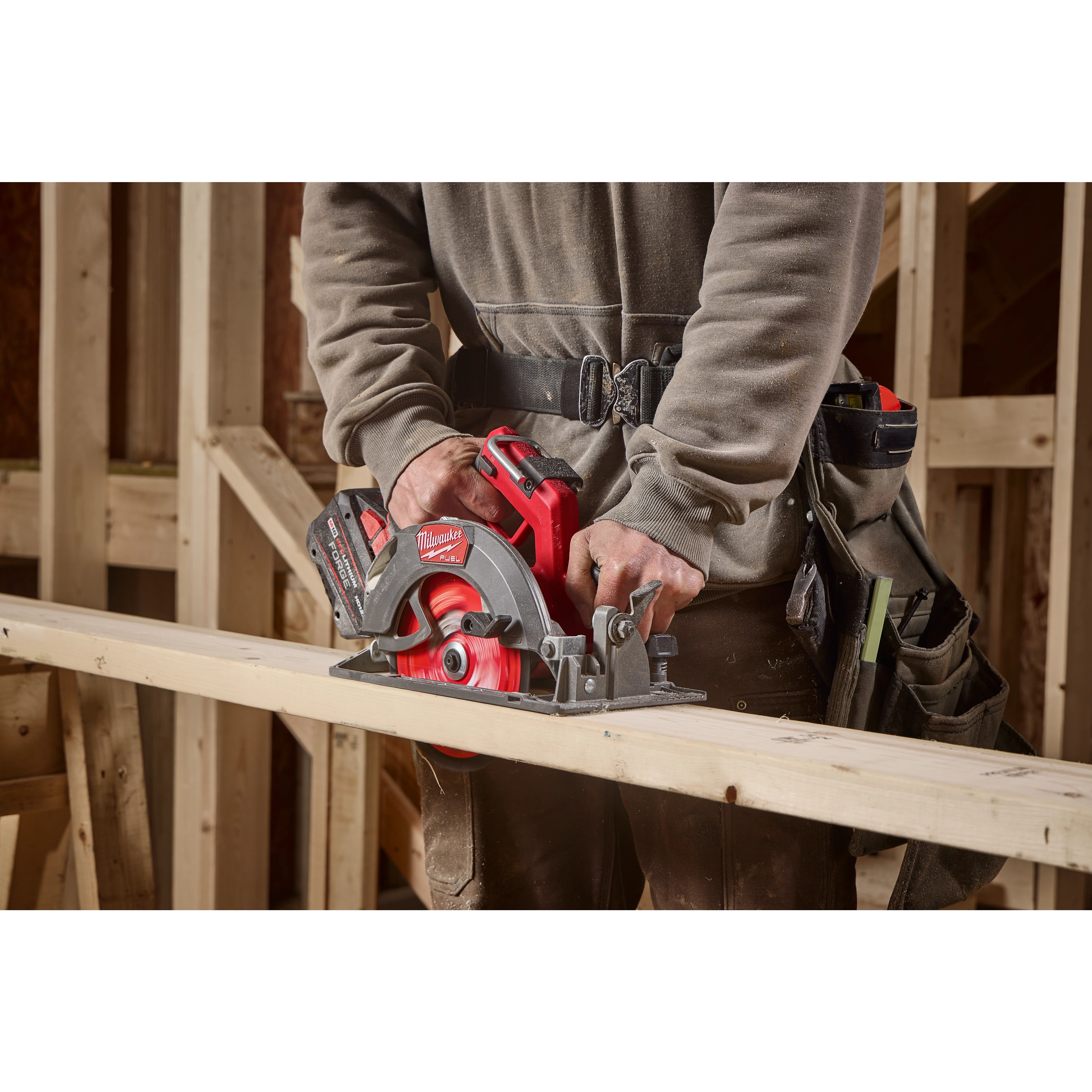 A person operates a Milwaukee circular saw to cut a wooden plank. The saw is red and grey, with "Milwaukee" visible on the side. The individual is dressed in a grey hoodie, brown pants, and a tool belt in a wood construction setting.