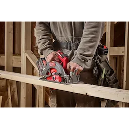 A person operates a Milwaukee circular saw to cut a wooden plank. The saw is red and grey, with "Milwaukee" visible on the side. The individual is dressed in a grey hoodie, brown pants, and a tool belt in a wood construction setting.