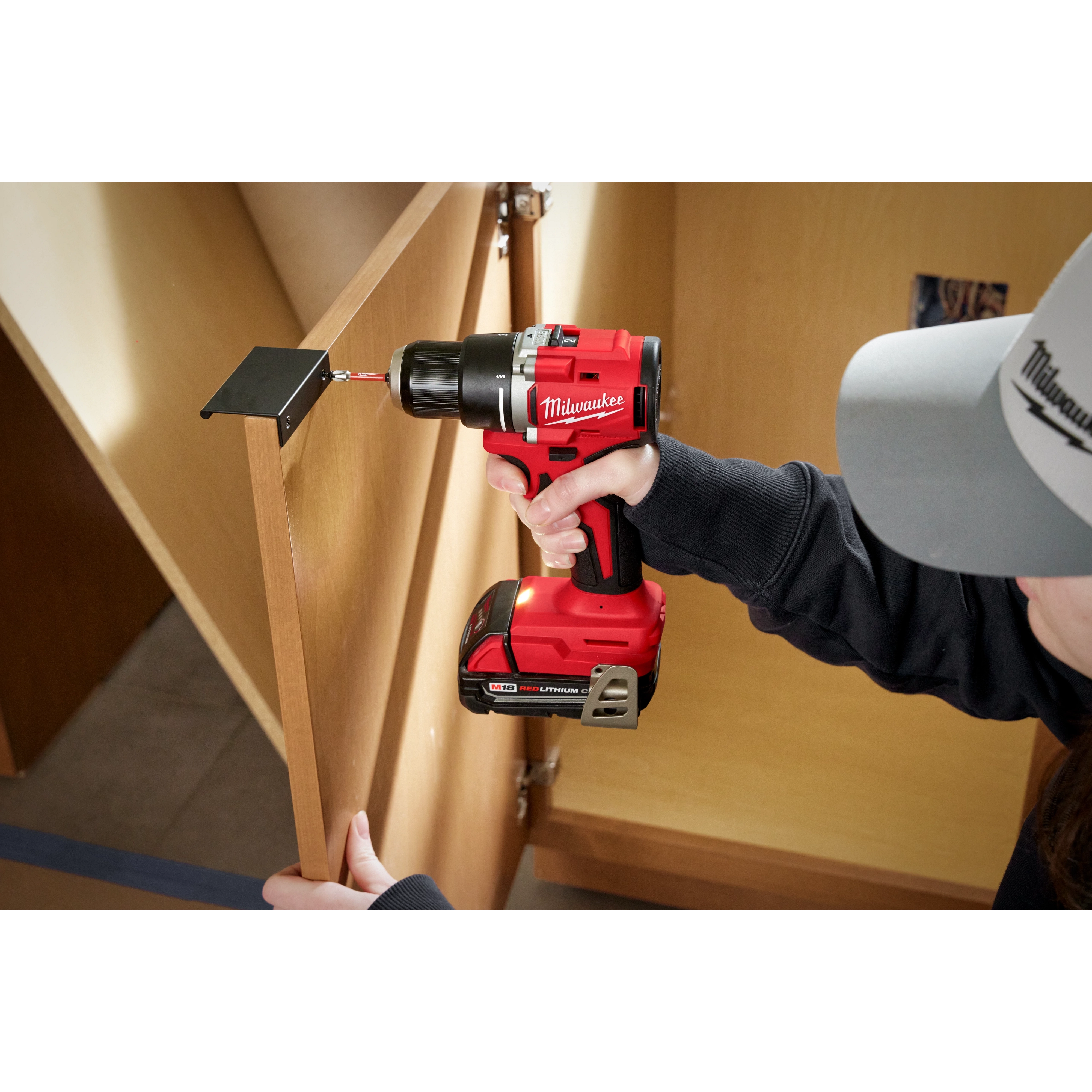 A person uses the M18 Compact Brushless 1/2" Drill/Driver to install a hinge on a wooden cabinet door indoors.
