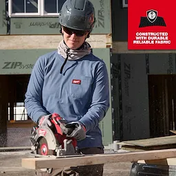 A worker wearing a Women's GRIDIRON™ Pocket T-Shirt - Long Sleeve in blue uses a circular saw on a wooden plank at a construction site. A red label indicates the shirt is constructed with durable, reliable fabric.