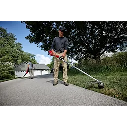 A person uses the M18 FUEL™ Power Head w/ QUIK-LOK™ to trim grass along a driveway. Another person in the background uses a blower. They are working near a house with a garage and trees around.
