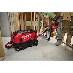 A person operates a red M18 FUEL™ PACKOUT™ 9 Gallon Dual-Battery Dust Extractor w/ VACLINK™ in a construction setting. The dust extractor is connected to a cutting tool, collecting dust and debris. The person is standing beside wooden studs and plumbing pipes in an unfinished room.