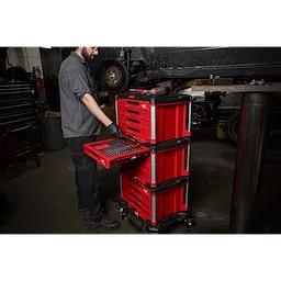 A mechanic is using a 366pc. Master Mechanics Hand Tool Set with PACKOUT™ Drawers and Dolly in a garage. The red tool chest is on wheels and has multiple drawers filled with tools.